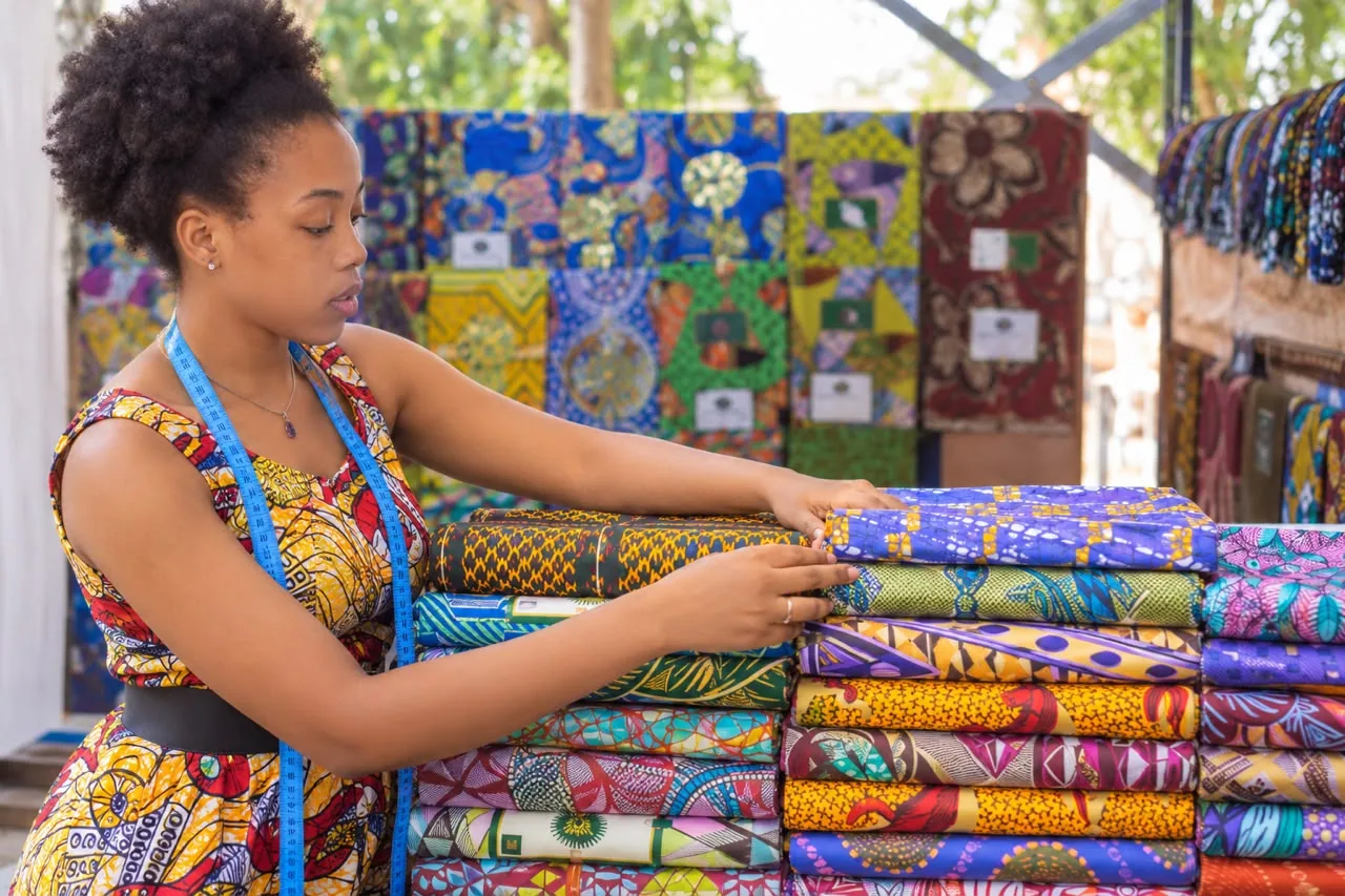 Tailor presenting neatly folded stacks of wax-print fabric at her market stall